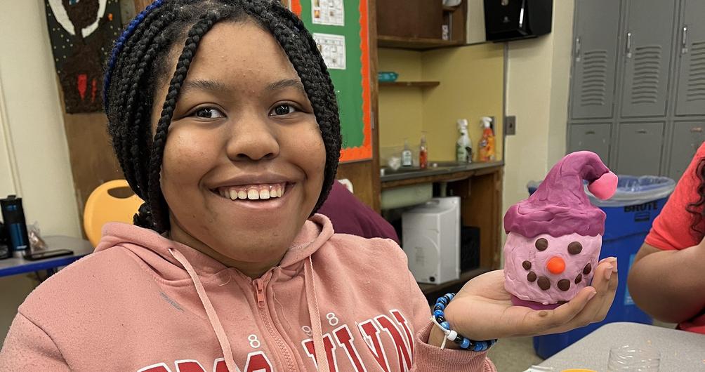 Girl smiling while holding a colorful clay cupcake model in a classroom setting.
