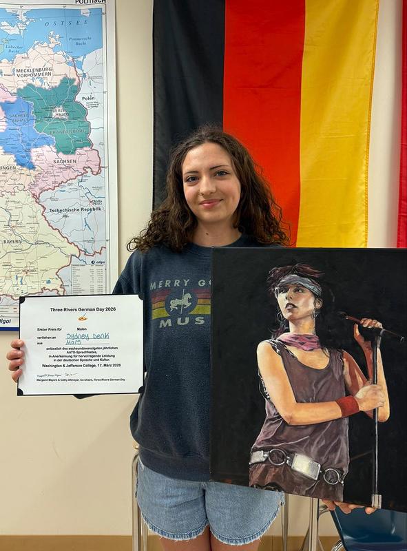 A student holds a certificate and a painted artwork, with a map and flag in the background.