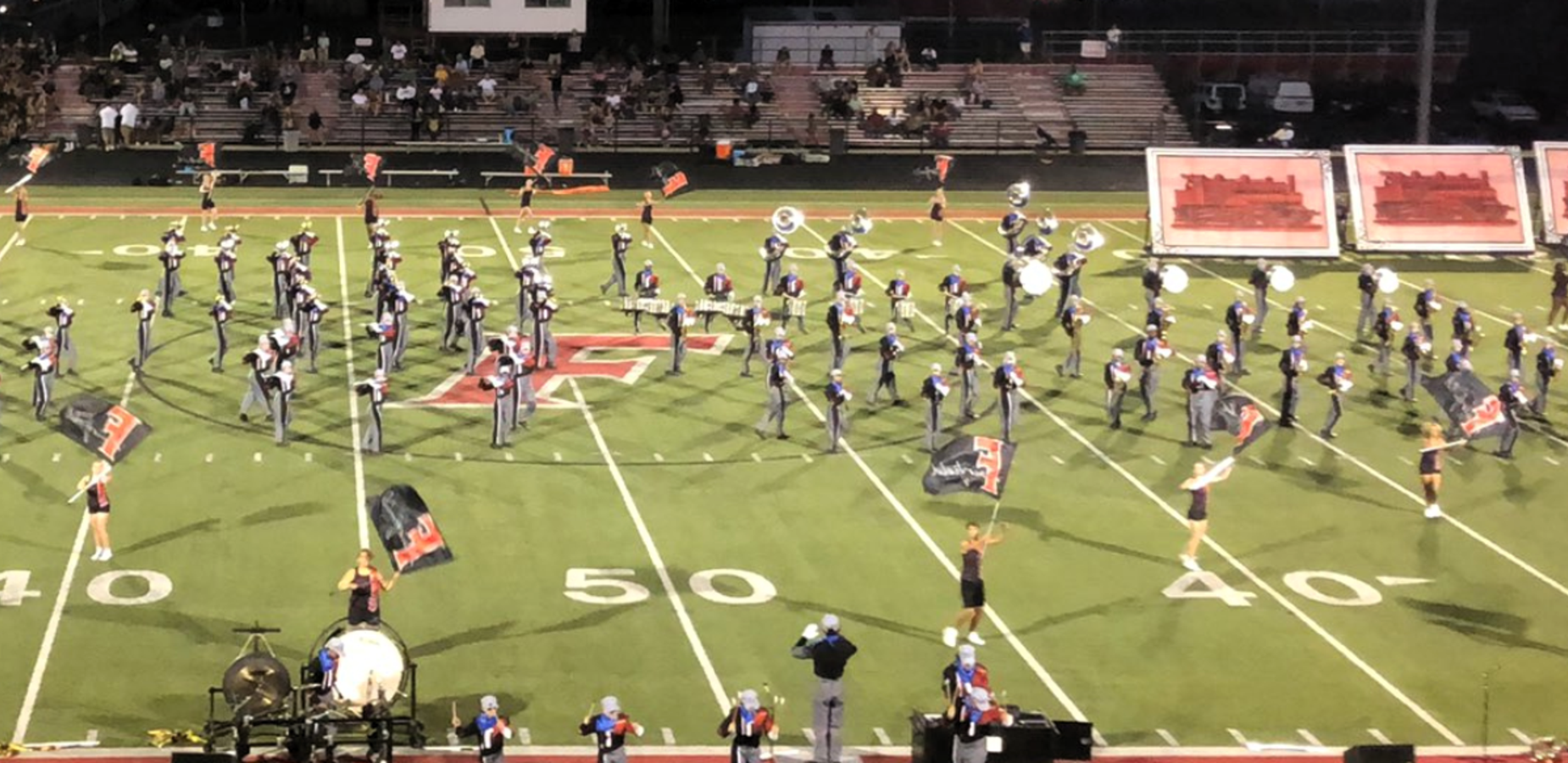 A marching band performing on a football field at night, with flags and instruments.
