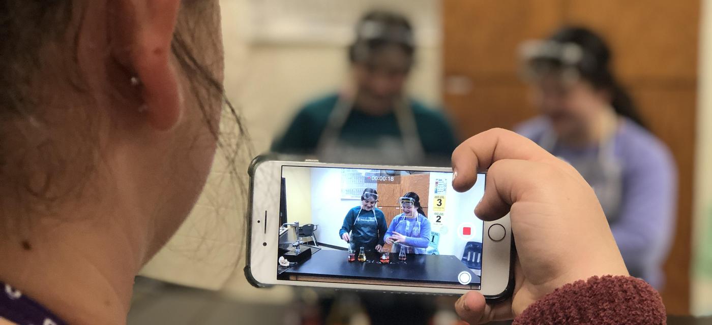 girl using camera phone to record 2 other girls in protective eye wear and aprons work with beakers of fluid