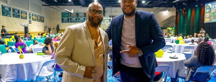 Two men in suits posing together at a festive event with tables and attendees in the background.