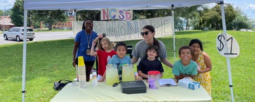 A group of kids at a lemonade stand with signs and drinks.