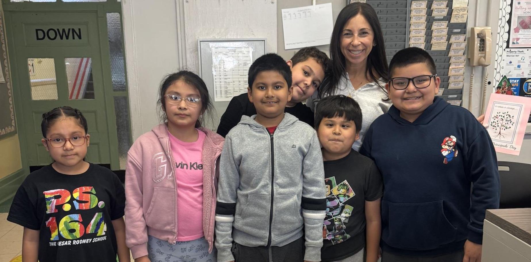 Group of seven children and an adult standing together in a classroom setting.