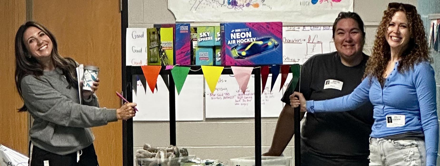 Three people stand next to a cart filled with various supplies in a classroom.