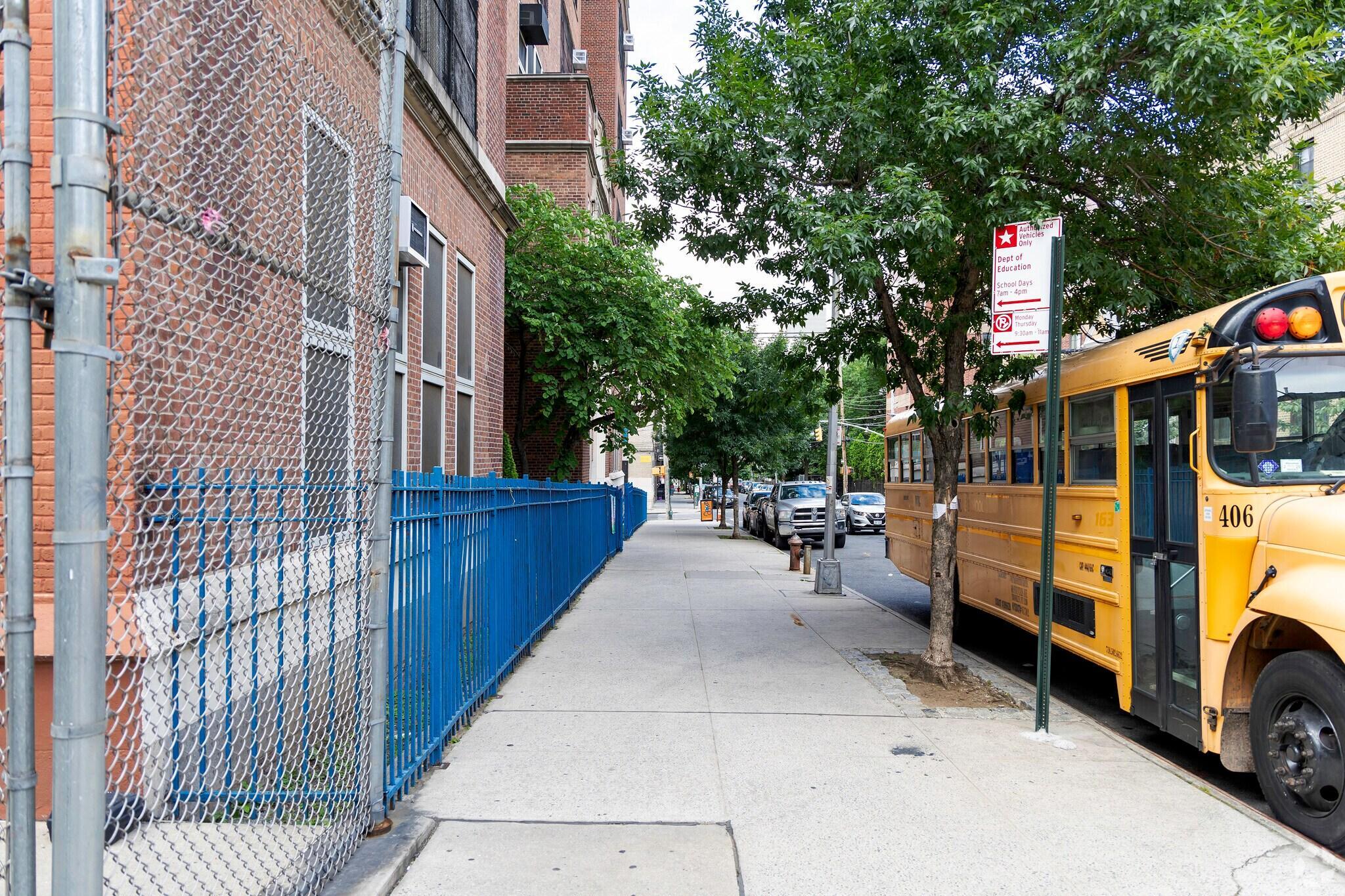 Sidewalk view of an urban school with a yellow school bus parked beside it. A chain-link fence and green trees line the quiet street.