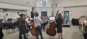 Several young musicians stand with double bass instruments in a classroom at Harmony Magnet Academy, practicing together as part of the Porterville Strings Summer Camp. Music stands and percussion instruments can be seen in the background, indicating an active and well-equipped music room.