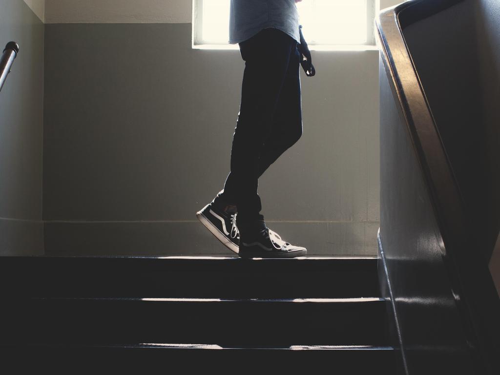 A person wearing black sneakers ascends a staircase, partially illuminated by a window.