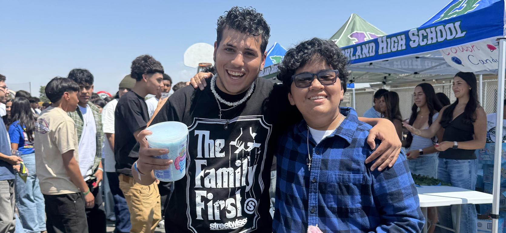 Two friends pose together at a lively outdoor event, smiling and holding drinks.