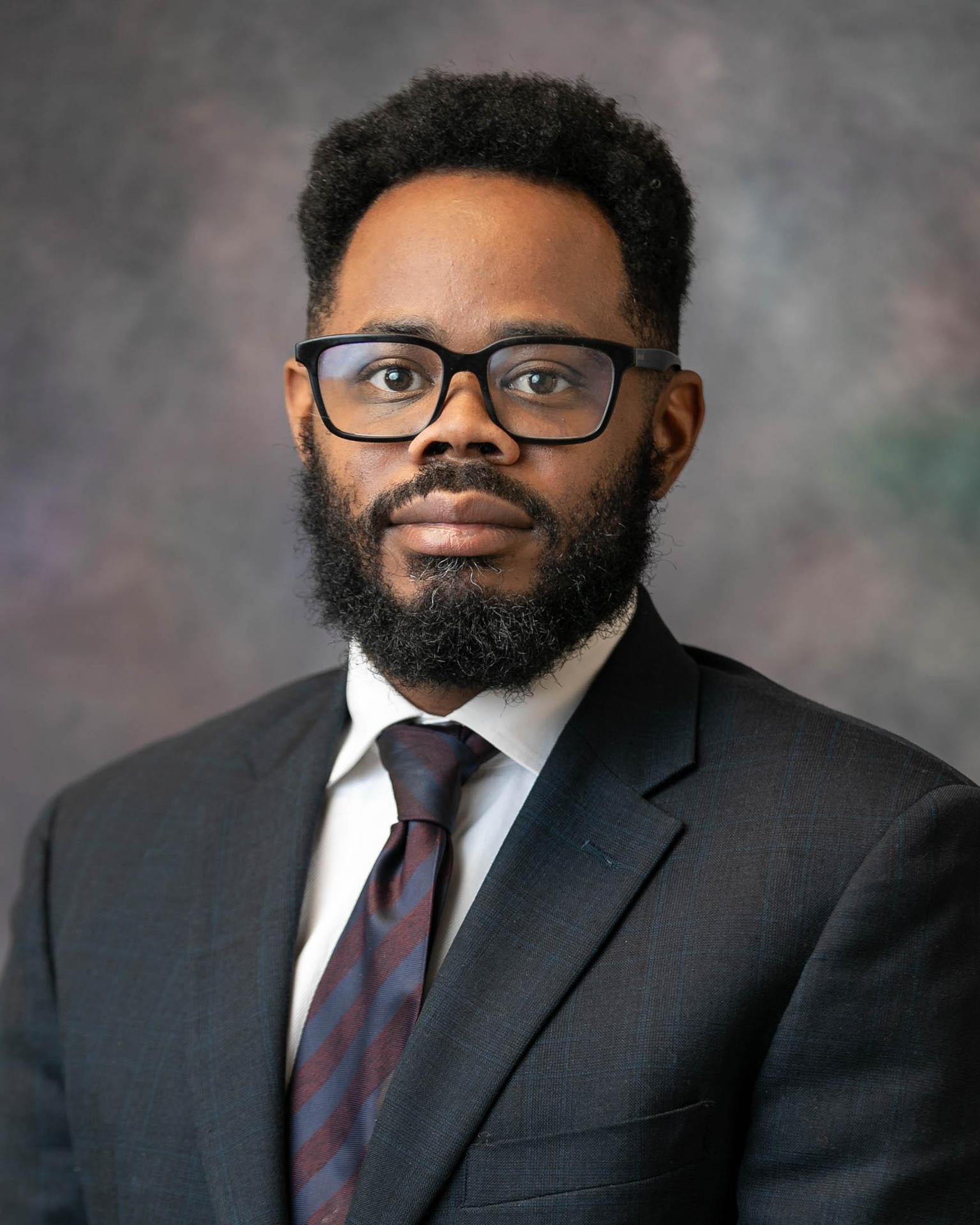 Headshot of a man in a black jacket with a red tie and gray beard