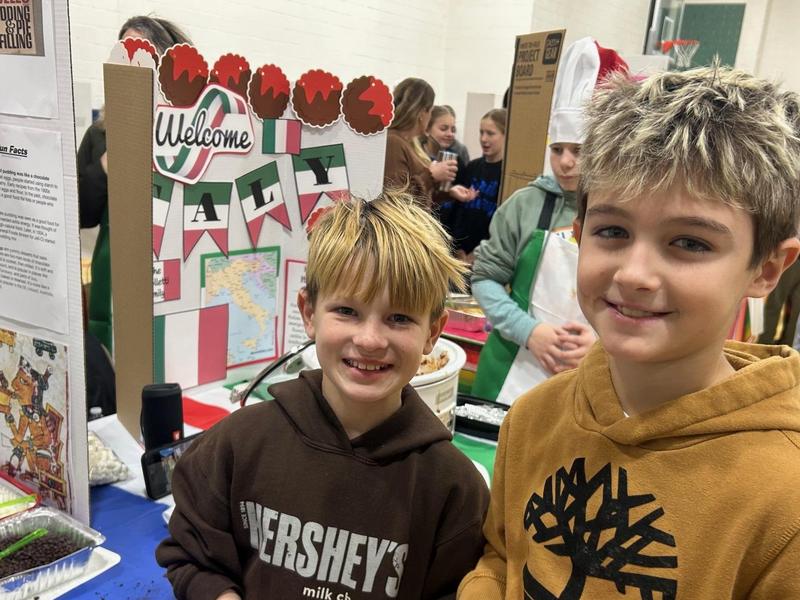Two boys smile in front of a 'Welcome to Italy' display with flags and a map.