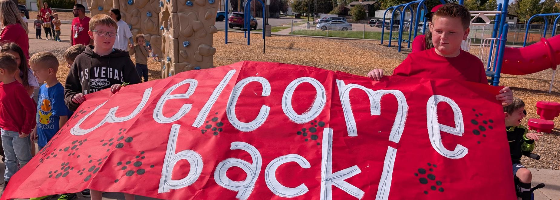 Children holding a large red banner that says 'welcome back!' at a playground.