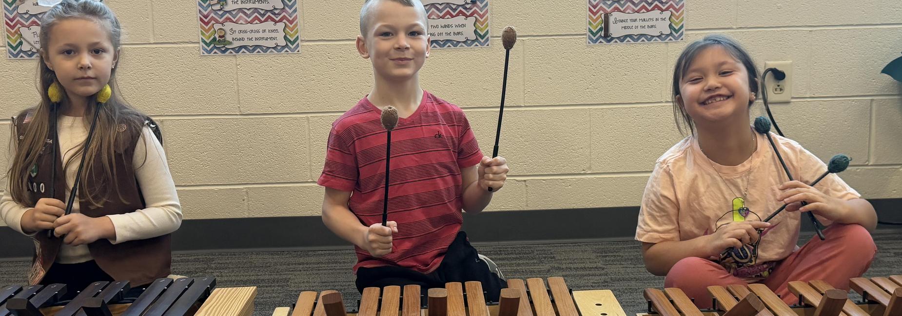 Three children playing wooden xylophones with mallets, smiling and enjoying music.