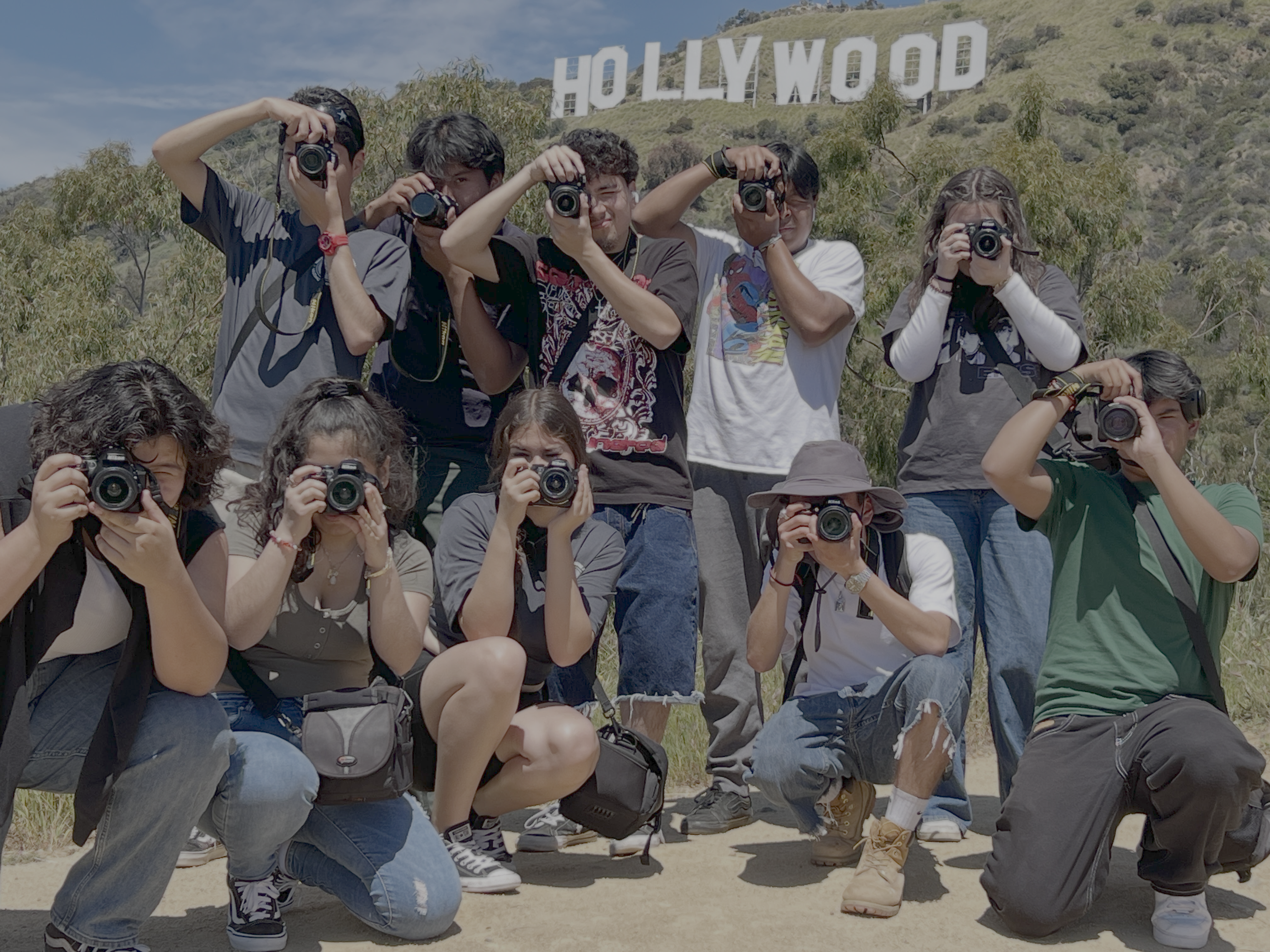 A group of students pose with cameras in front of the Hollywood Sign during a photography outing. Several students hold cameras up to their faces while others kneel or stand together on a sunny hillside.