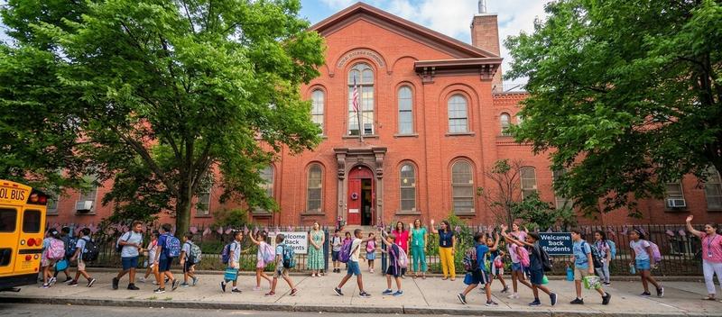 Children and teachers outside the red school building coming to school.