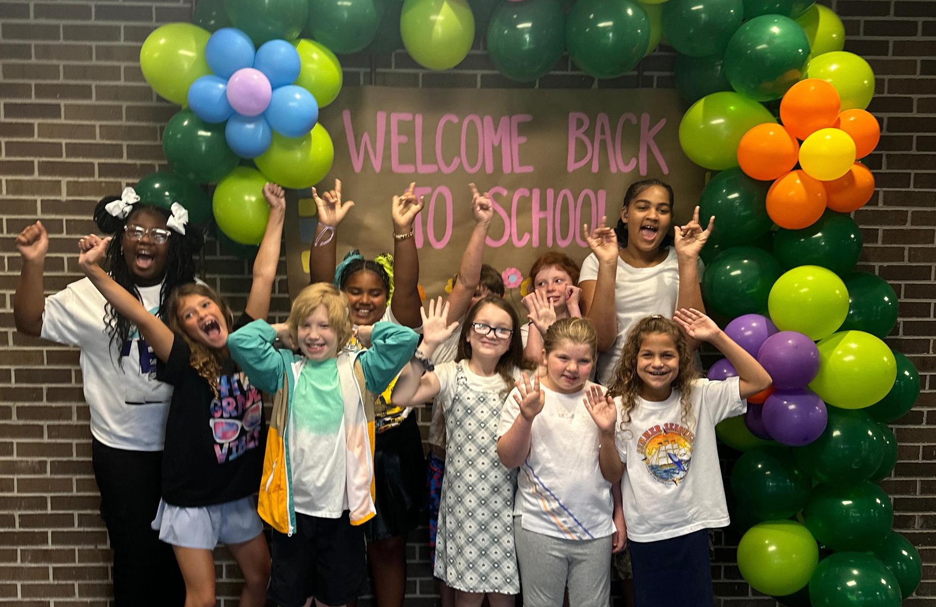 Group of children cheering under a colorful balloon arch at a school welcome event.
