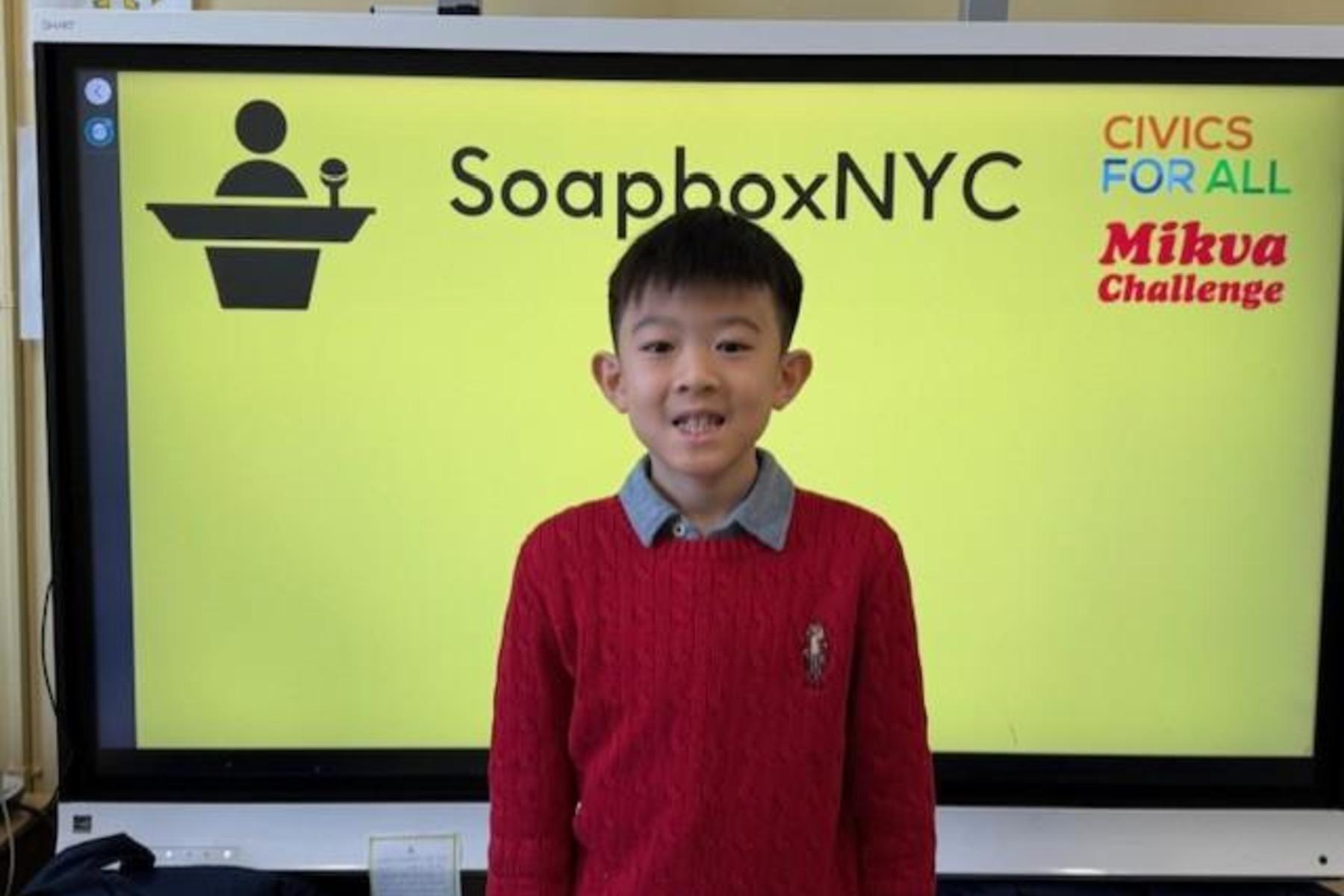 Boy in a red sweater smiles in front of a bright yellow background with text about SoapboxNYC.