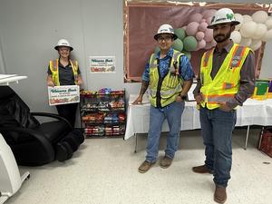 Three construction workers in yellow vests and white hardhats standing near snack basket