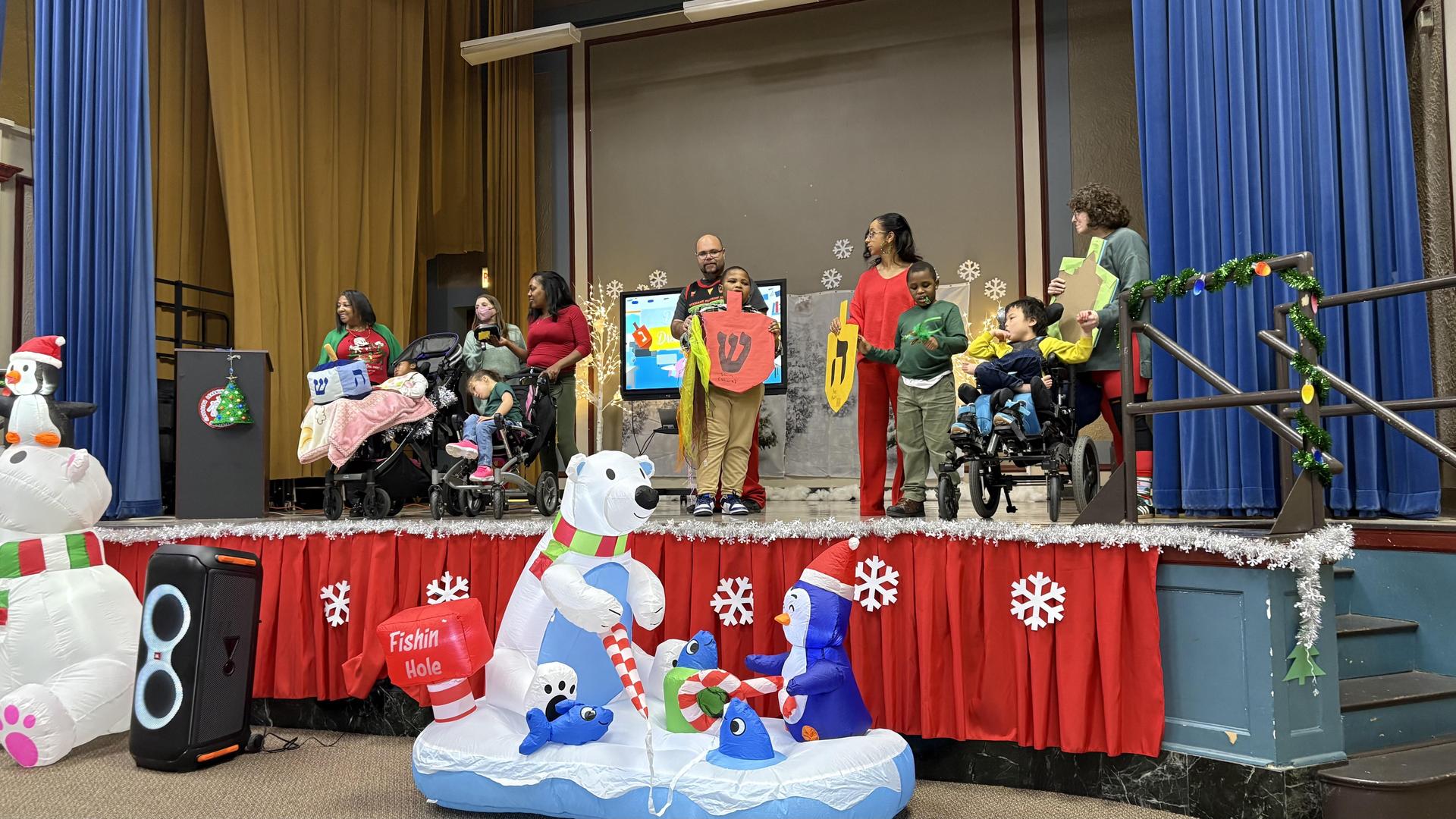 Children and adults on stage in festive attire celebrating with decorations and an inflatable polar bear.