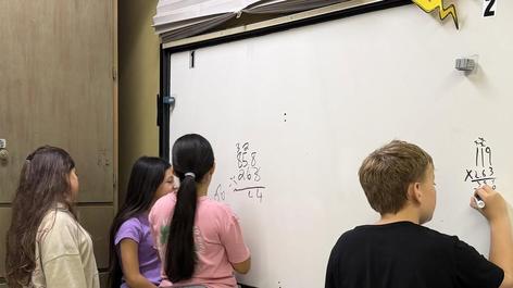 Four children solving math problems on a whiteboard in a classroom.