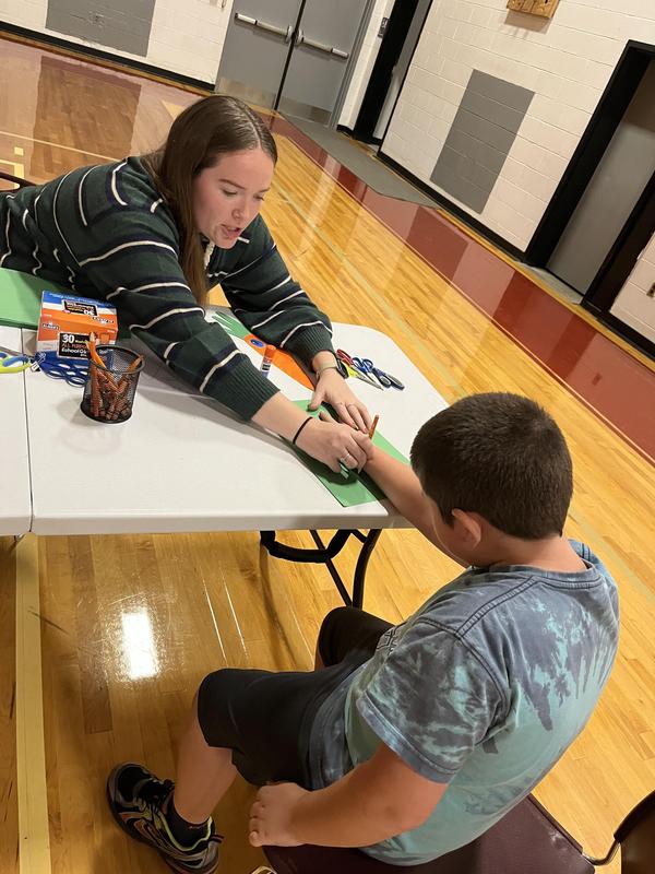 Teen helping a boy with his art project at a table.