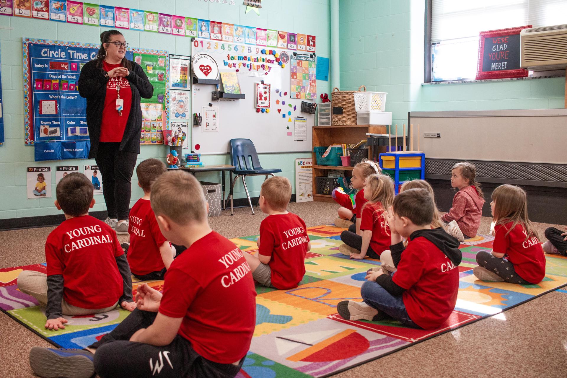 Preschool students listening to their teacher at the front of the classroom.