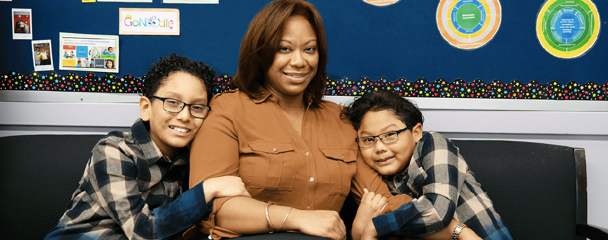 A woman sits with two boys, smiling in a classroom setting.
