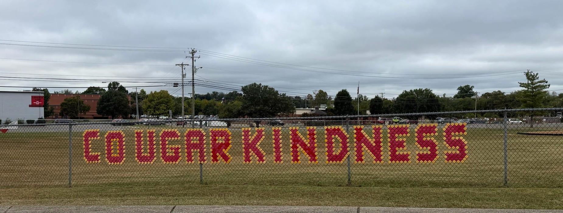 Colorful letters spelling 'COUGAR KINDNESS' on a fence with a cloudy sky.