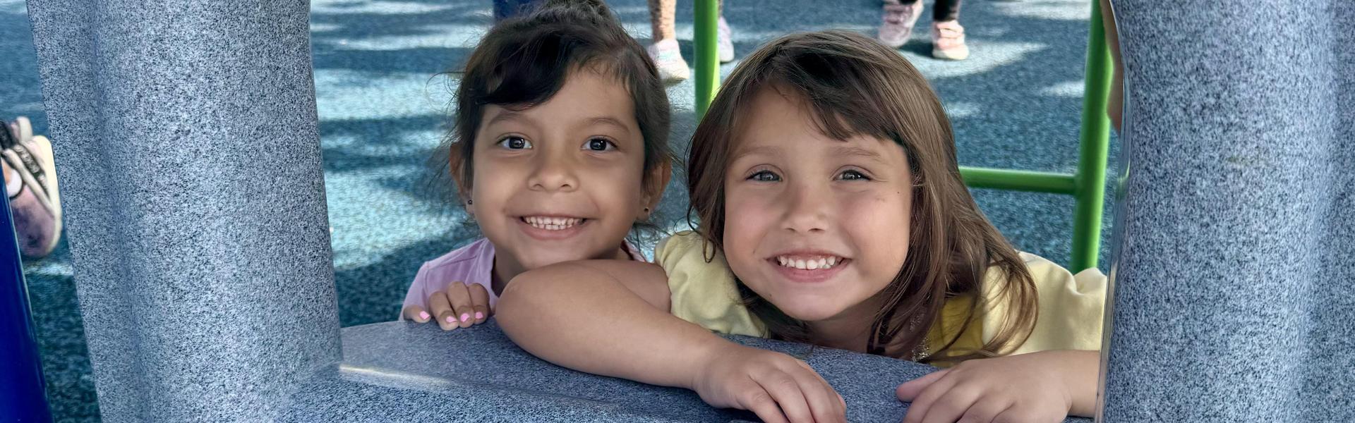 Two smiling girls peeking through playground equipment.