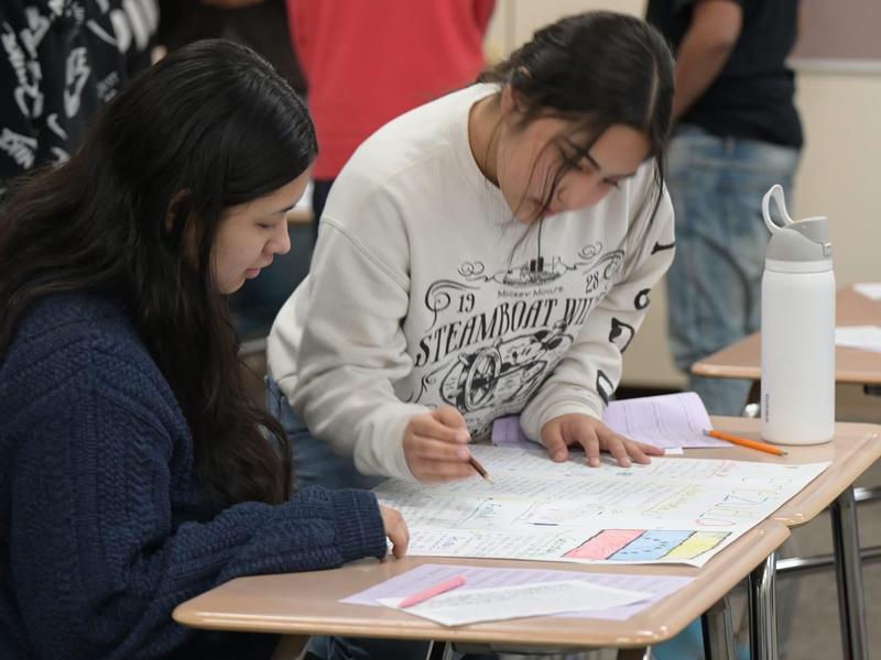 Two students working collaboratively at a desk with papers and a water bottle.