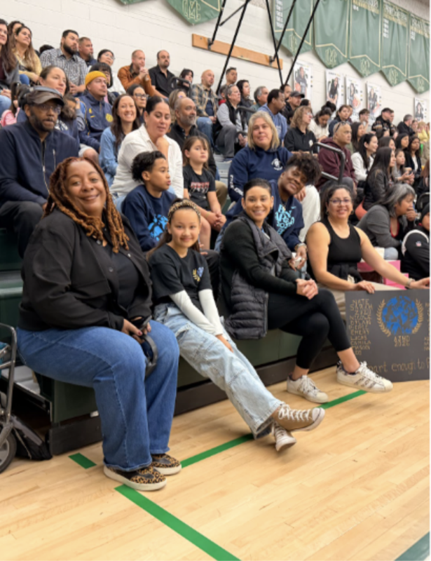Parents, alternates, and family members cheer on the teams. A parent can also be seen holding a sign that matches the team shirts.