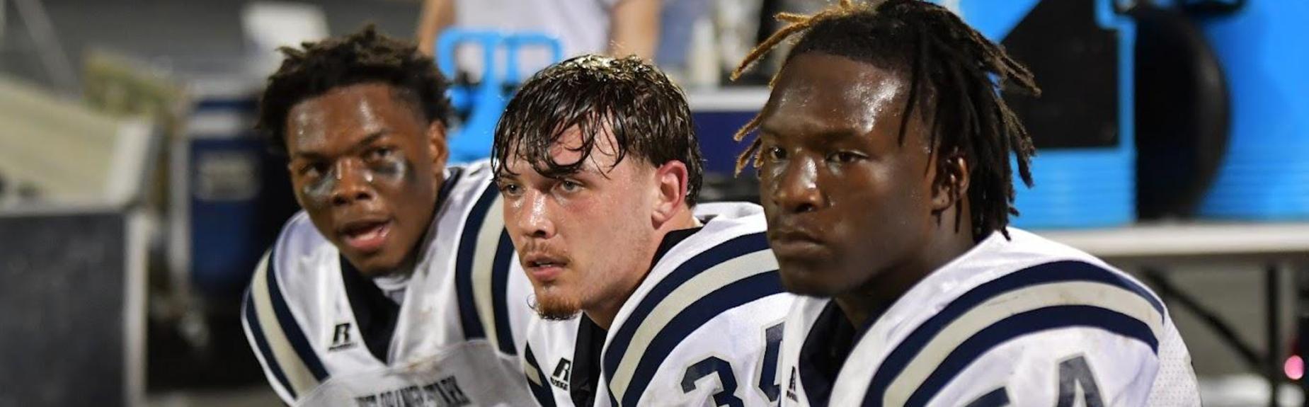 Three football players on the sidelines, looking focused and discussing the game.