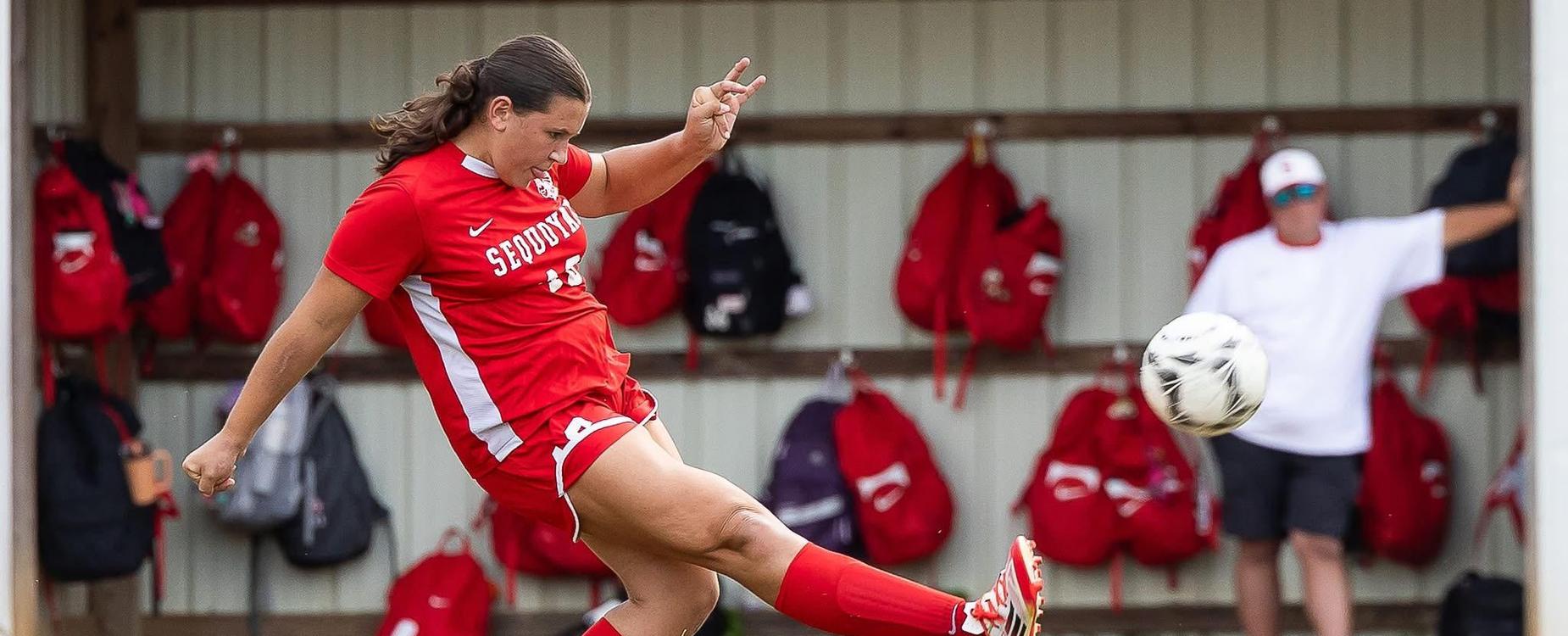 A soccer player in red kicks a ball during a game, with bags hanging in the background.