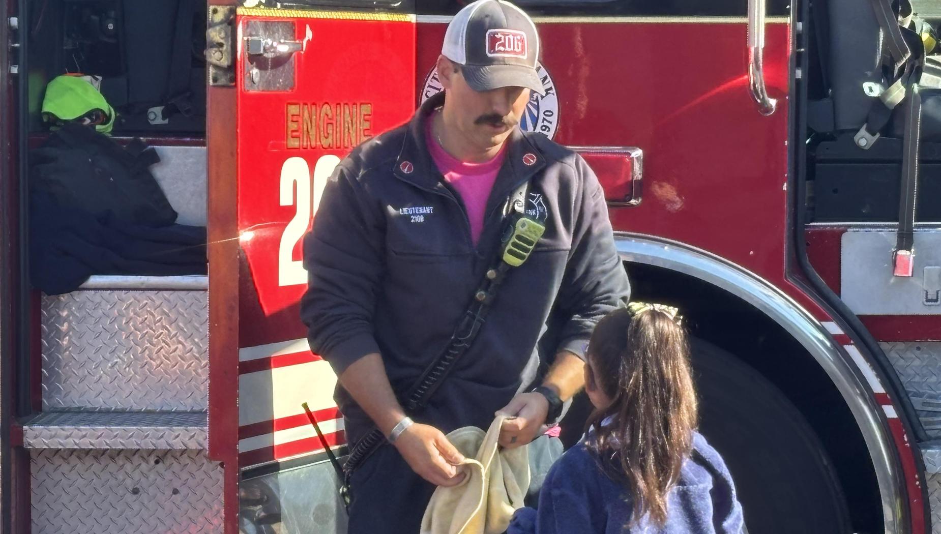 Firefighter talking to a child near a fire truck, wearing uniform and cap.