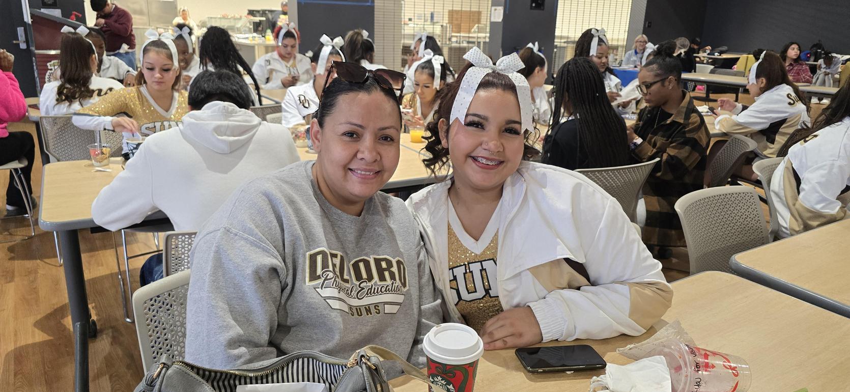 Two women smiling together at a gathering in a busy cafeteria.