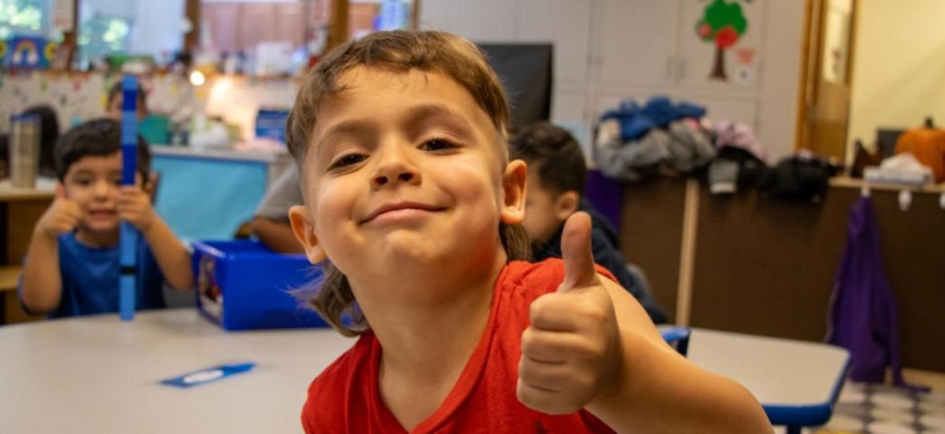 Child giving a thumbs-up in a classroom with other kids in the background.