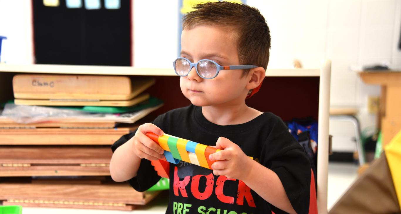 A young child wearing glasses holds a colorful toy in a classroom setting.