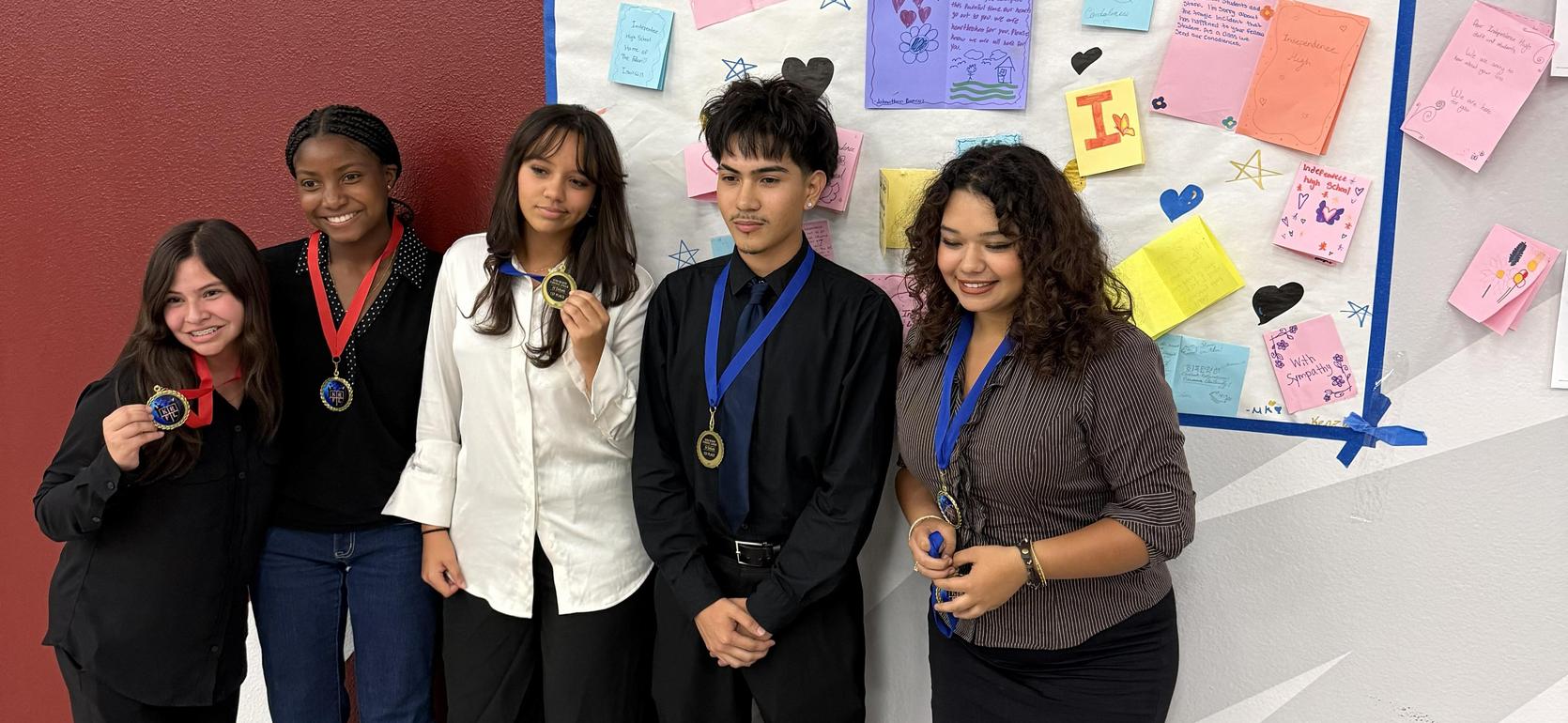 Five students holding medals in front of a colorful bulletin board.