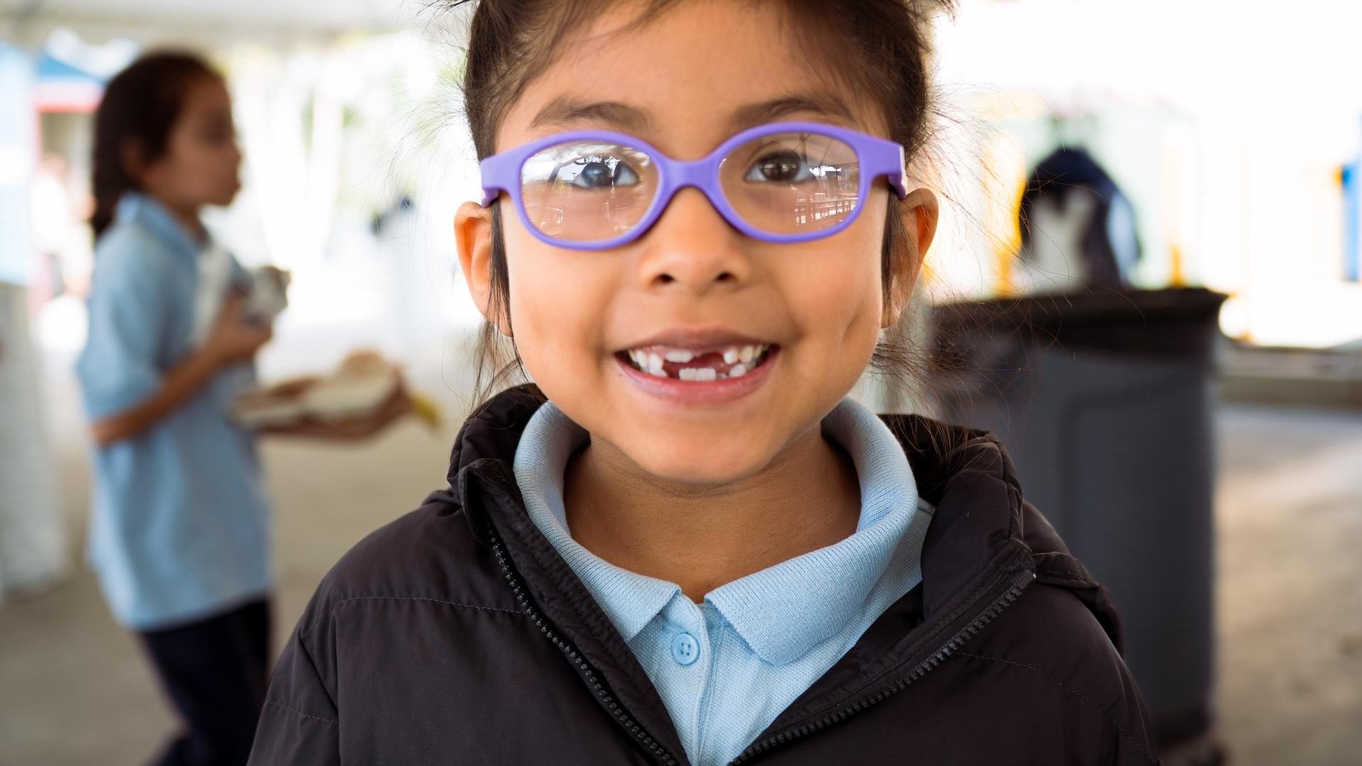 A girl with glasses smiles widely, revealing her missing teeth.