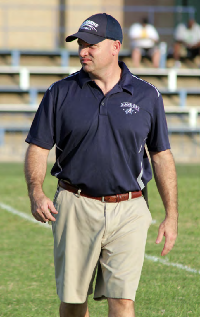 Texas School for the Deaf (TSD) Coach Chris Hamilton stands on the football field, looking focused and ready for action. Dressed in a navy blue polo shirt with the TSD Rangers logo and khaki shorts, he exudes a determined and confident presence. He wears a matching TSD Rangers cap as he observes the field, with blurred bleachers and spectators in the background, emphasizing the setting of a school sports event.