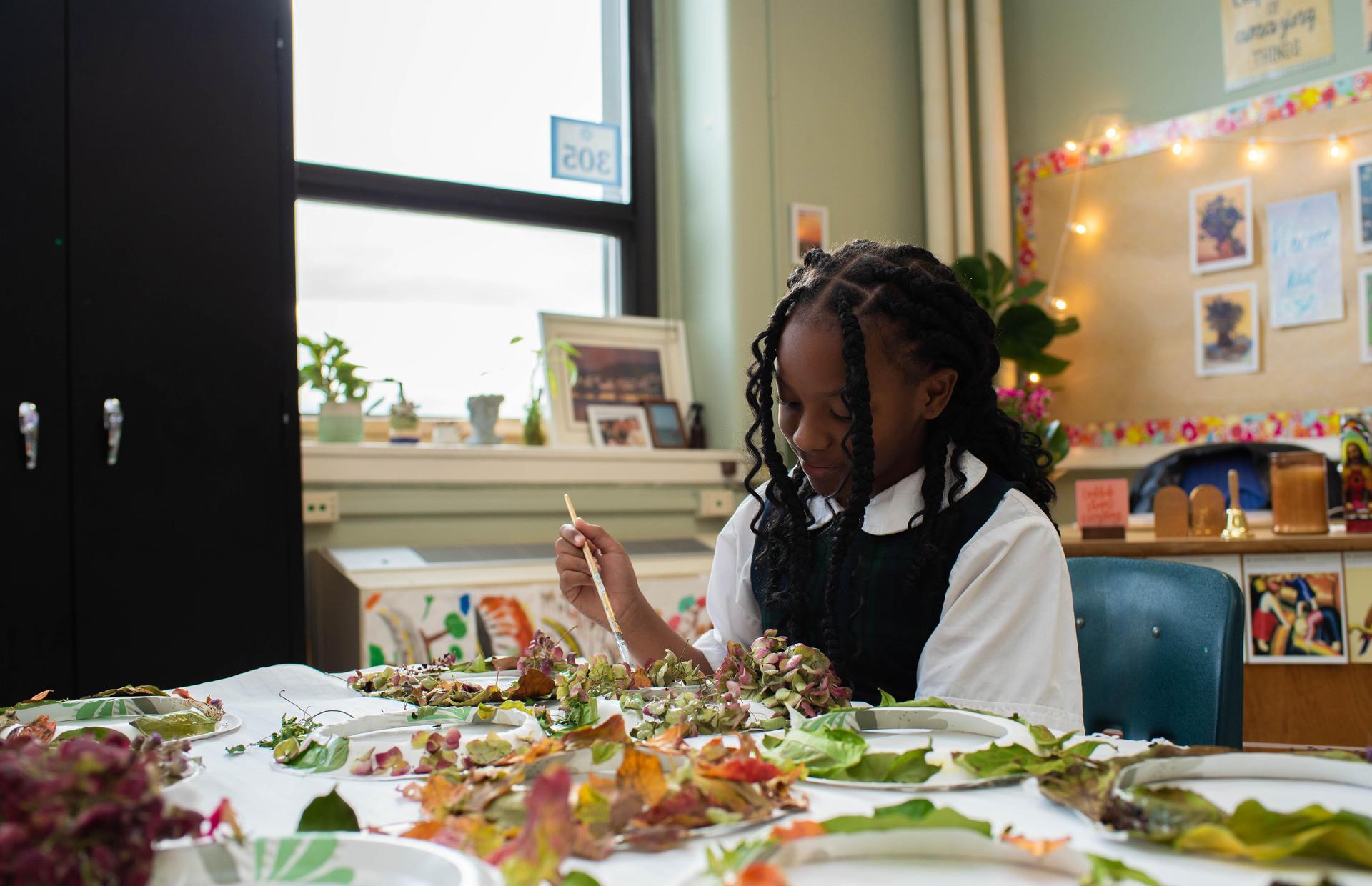 A girl focuses on crafting with leaves at a table, surrounded by plants and art supplies.