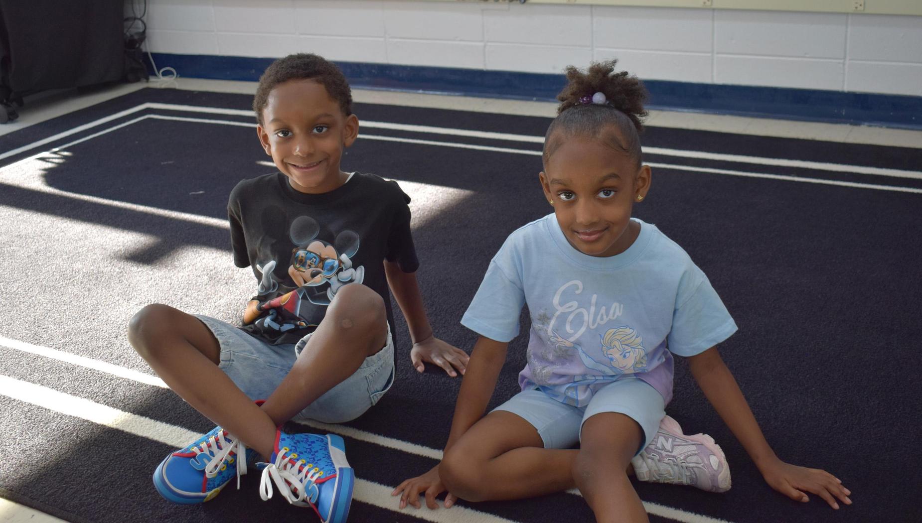 Two children sit on a carpet in a classroom, smiling and posing for the camera.