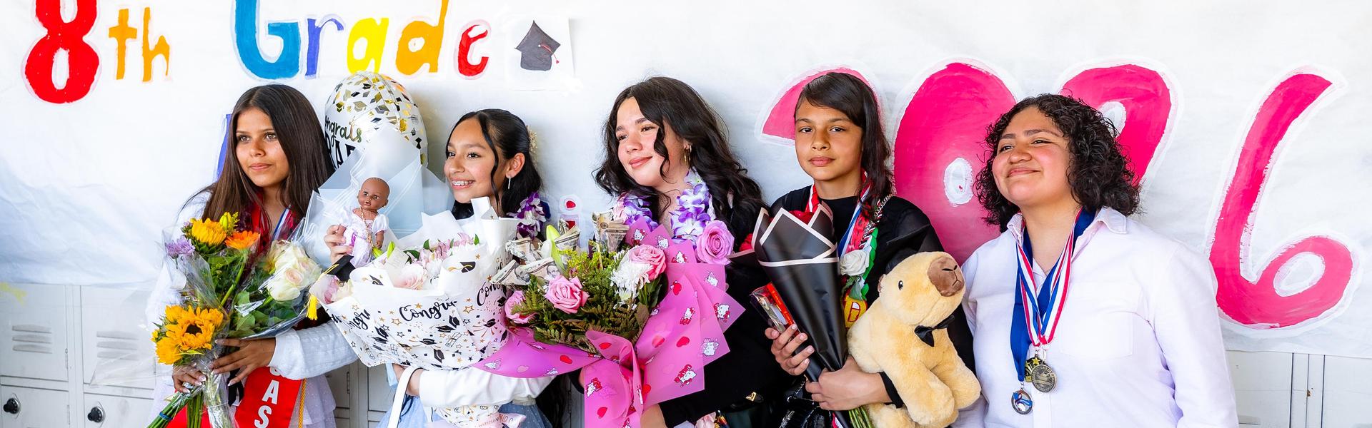 Five girls posing with flowers and awards in front of a colorful graduation banner.