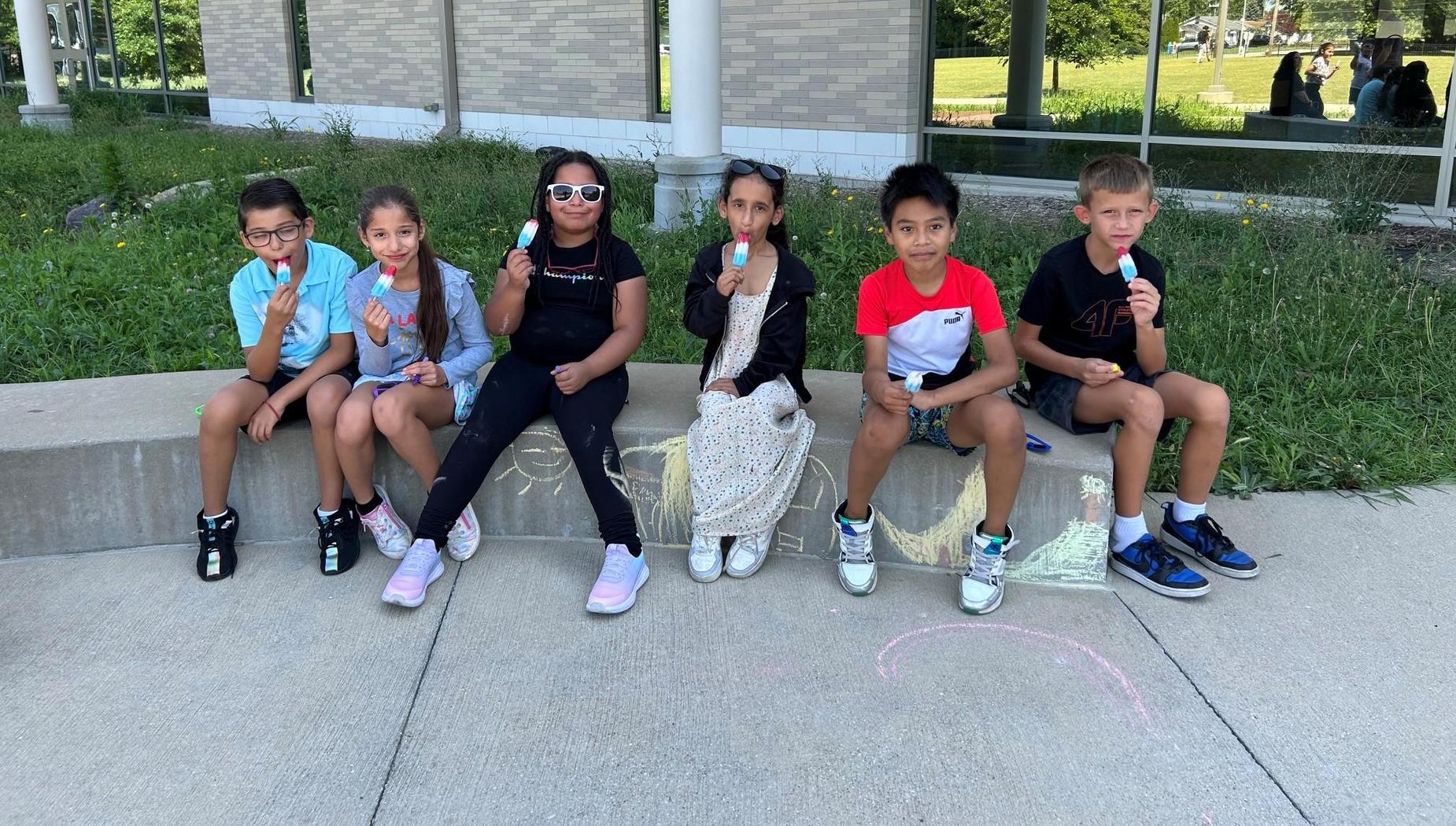 Group of six children sitting outside enjoying popsicles.