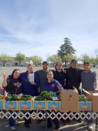 Group of six people smiling behind boxes of vegetables outdoors.