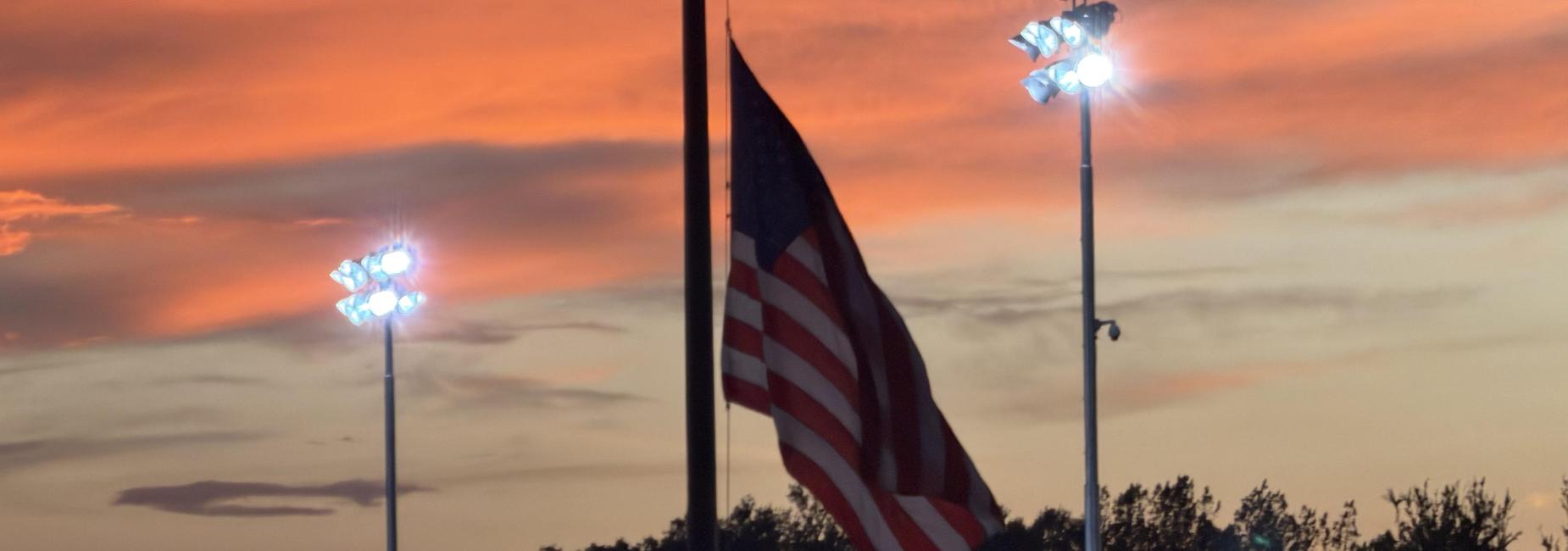 American flag silhouetted against a sunset sky with stadium lights.
