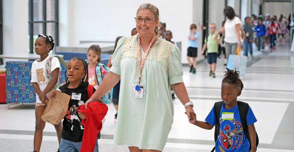 Two students hold hands with a teacher while walking down the hall.
