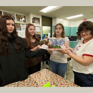 Four students presenting a decorated dessert in a classroom setting.