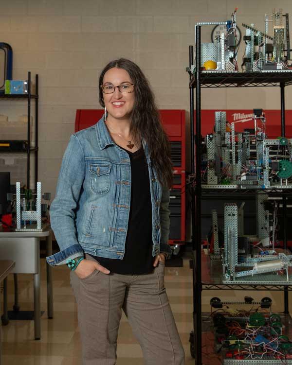Woman with glasses smiling in a workshop with robotics behind her.
