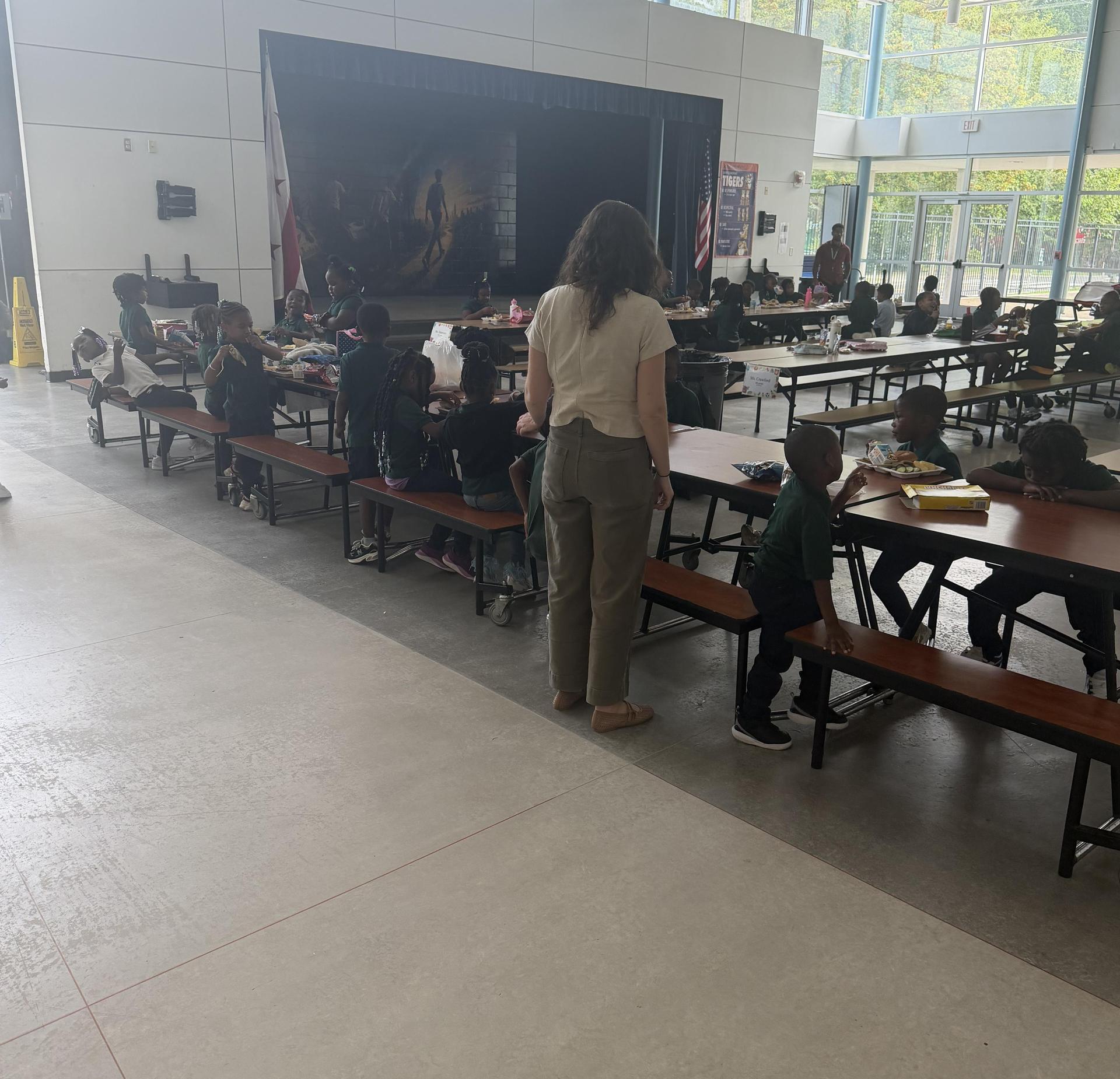 Students seated at long tables in a cafeteria, with an adult overseeing them.