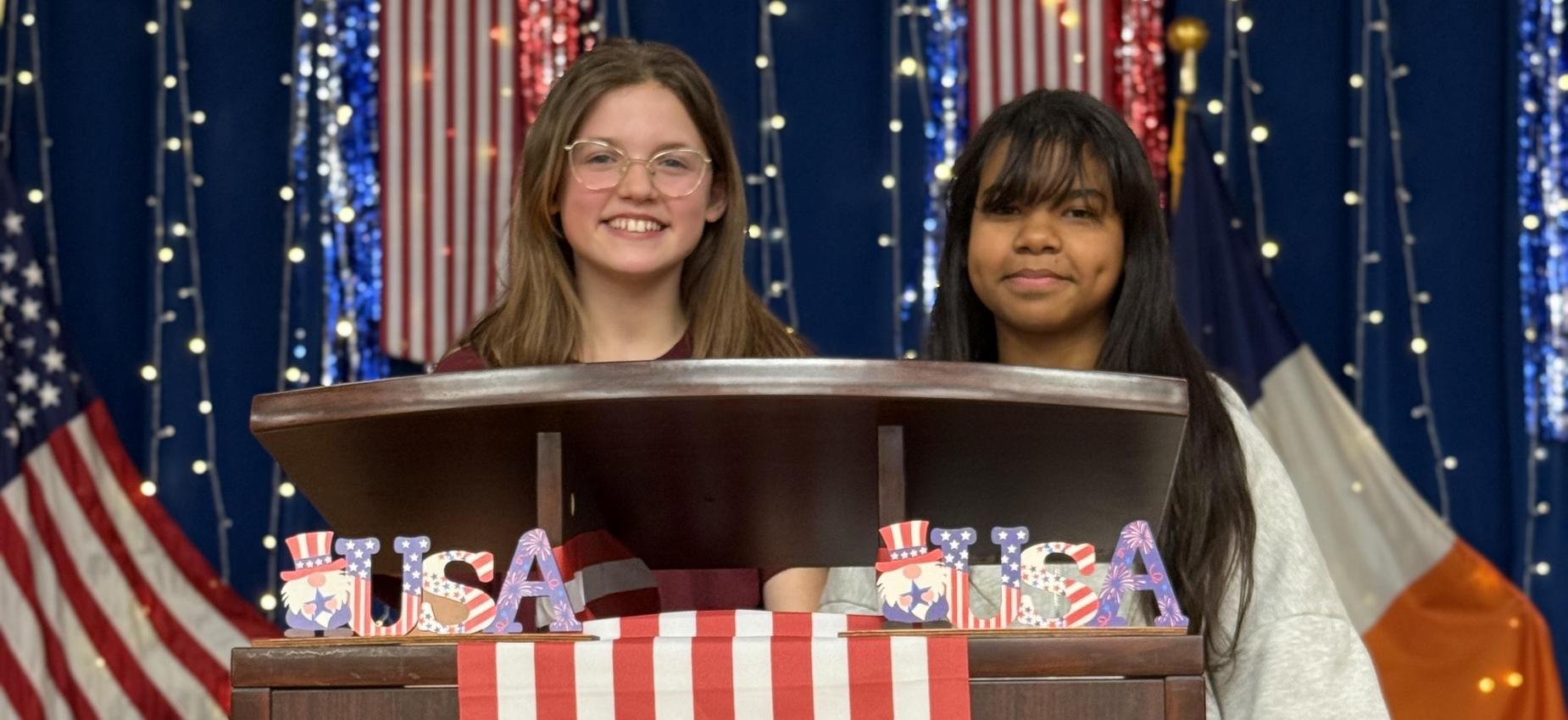 Two girls in front of a podium with an American flag.